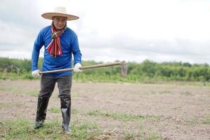 asiatischer Bauer trägt Hut, blaues Hemd, hält eine Hacke, um Erde in seinem Garten zu graben. konzept, landwirtschaftsberuf, ökologischer landbau, verwenden sie manuelles werkzeug, um unkraut loszuwerden. foto