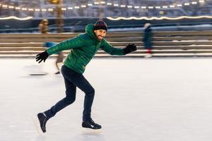 Aufnahme eines fröhlichen Mannes hat Spaß im Freien, läuft Schlittschuhe auf dem Eisring, genießt diese Winteraktivität, bewegt sich aktiv, versucht, das Gleichgewicht zu halten, ist gut trainiert, schaut mit glücklichem Ausdruck in die Kamera foto