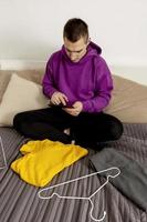 Mann mit violettem Hoodie mit digitalem Tablet macht ein Foto seiner alten Kleidung, um sie online zu verkaufen. Verkauf auf der Website, E-Commerce. Wiederverwendung, Second-Hand-Konzept. bewusster Konsument, nachhaltiger Lebensstil