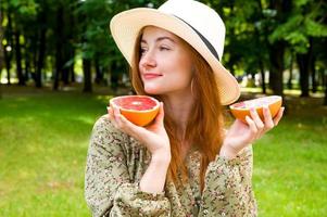 junge glückliche rothaarige Frau in einem Hut, der Grapefruit hält. sommerporträtkonzept. Obst und Vitamine foto