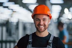 lächelnder und glücklicher mitarbeiter. Porträt des Industriearbeiters drinnen in der Fabrik. junger Techniker mit orangefarbenem Schutzhelm foto