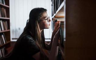 Suche nach richtigen Informationen. gegen Fenster. Studentin ist in der Bibliothek, die voller Bücher ist. Konzeption von Bildung foto