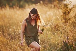 Schönes Mädchen geht mit hohem Gras durch das Feld und sammelt Blumen. erstaunliches Sonnenlicht foto