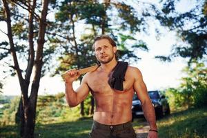 Porträt des Holzfällers mit Axt in der Hand hautnah. Ein hübscher, hemdloser Mann mit muskulösem Körpertyp ist tagsüber im Wald foto