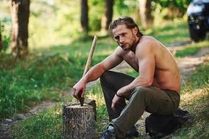 hält Holzaxt. Ein hübscher, hemdloser Mann mit muskulösem Körpertyp ist tagsüber im Wald foto
