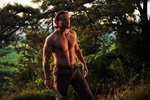 bereit für die Arbeit. selbstbewusster Holzfäller. Ein hübscher, hemdloser Mann mit muskulösem Körpertyp ist tagsüber im Wald foto