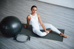 Sitzen auf Rollmatte. junge sportliche frau in weißer kleidung mit schlankem körpertyp im gesunden zentrum macht eine pause. schwarzer Fitnessball foto