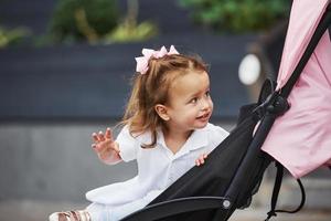 Porträt eines fröhlichen kleinen Mädchens, das im rosafarbenen Kinderwagen sitzt, während es in der Stadt spazieren geht foto