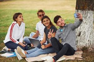 neben dem Baum sitzen und Selfie machen. Gruppe junger Studenten in Freizeitkleidung auf grünem Gras tagsüber foto