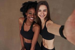 Selfie ankommend. Zwei multiethnische Freundinnen stehen im Studio mit braunem Hintergrund foto