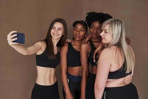 Selfies machen. Gruppe multiethnischer Frauen, die im Studio vor braunem Hintergrund stehen foto