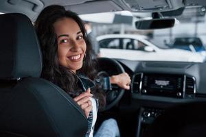 rückwärts bewegen. ute mädchen mit schwarzen haaren, die ihr brandneues teures auto im autosalon ausprobieren foto