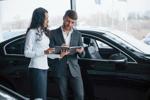 müssen dieses Dokument unterschreiben. Kundin und moderner stylischer bärtiger Geschäftsmann im Autosalon foto