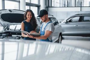 lesen Sie dies sorgfältig durch. Frau im Autosalon mit Angestellter in blauer Uniform, die ihr repariertes Auto zurücknimmt foto