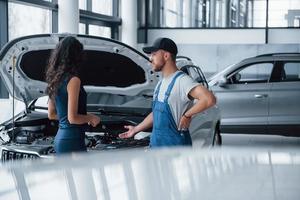 einige Details über Unfall. Frau im Autosalon mit Angestellter in blauer Uniform, die ihr repariertes Auto zurücknimmt foto