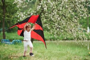 positives weibliches kind, das draußen mit rot-schwarz gefärbtem drachen in den händen läuft foto