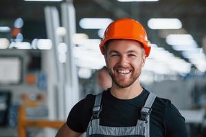 lächelnder und glücklicher mitarbeiter. Porträt des Industriearbeiters drinnen in der Fabrik. junger Techniker mit orangefarbenem Schutzhelm foto