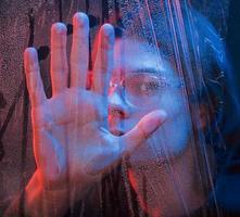 sag Hallo. Studioaufnahme im dunklen Studio mit Neonlicht. Porträt eines ernsthaften Mannes hinter dem nassen Glas foto