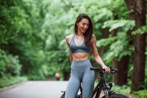 aufrichtiges Lächeln. Radfahrerin, die tagsüber mit dem Fahrrad auf einer asphaltierten Straße im Wald steht
