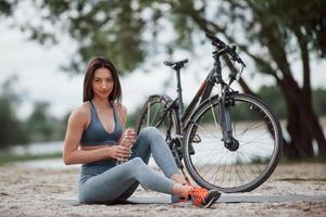 Wasserhaushalt des Menschen. Radfahrerin mit guter Körperform, die tagsüber neben ihrem Fahrrad am Strand sitzt foto