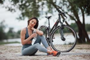 Wasser ist sehr wichtig. Radfahrerin mit guter Körperform, die tagsüber neben ihrem Fahrrad am Strand sitzt foto