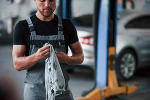 gewöhnlicher Arbeitstag. Mann in schwarzem Hemd und grauer Uniform steht in der Garage, nachdem er ein kaputtes Auto repariert hat foto