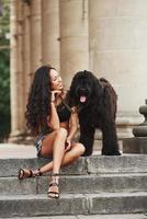 Hund steht in der Nähe seiner Besitzerin. schöne frau mit lockigem schwarzem haar hat tagsüber eine gute zeit in der stadt foto