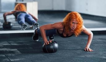 Liegestütze mit Ball machen. Sportliches rothaariges Mädchen hat tagsüber einen Fitnesstag im Fitnessstudio. muskulöser Körpertyp foto