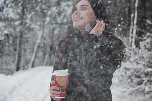 Schnee fällt vom Himmel. Mädchen in warmer Kleidung mit einer Tasse Kaffee spazieren im Winterwald foto