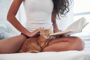 beschnittenes Foto. attraktive Blondine, die mit ihrem süßen Kätzchen auf dem weißen Bett ruht foto