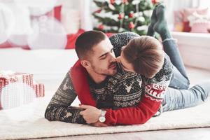 gib mir einen Kuss. Schönes junges Paar, das zur Neujahrszeit mit Geschenkboxen auf dem Boden des Wohnzimmers liegt foto