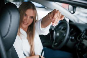 Glücklicher Besitzer mit Schlüsseln in der Hand. schönes blondes mädchen, das im neuen auto mit modernem schwarzem interieur sitzt foto