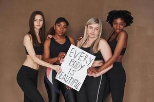 Jeder Körper ist schön, Zeichen. Gruppe multiethnischer Frauen, die im Studio vor braunem Hintergrund stehen foto