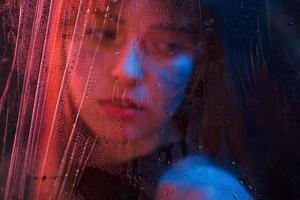 traurige Frau. Studioaufnahme im dunklen Studio mit Neonlicht. Porträt eines schönen Mädchens hinter nassem Glas foto