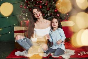 Fröhliche Mutter und Tochter, die hinter dem Weihnachtsbaum sitzen. süßes Porträt foto