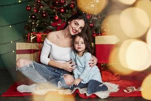 schöne junge Frau umarmt das Kind. Fröhliche Mutter und Tochter, die hinter dem Weihnachtsbaum sitzen. süßes Porträt foto