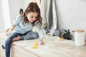 Hose ist schmutzig. Foto eines hübschen kleinen Mädchens, das auf dem Küchentisch sitzt und mit Mehl spielt