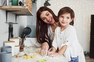 zur Seite schauen, während die Frau lächelt. glückliche tochter und mutter bereiten zusammen backwaren zu. kleiner Helfer in der Küche foto