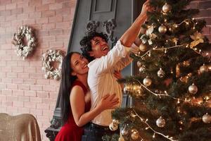 sieht aus wie lautes Lachen. Romantisches Paar schmückt Weihnachtsbaum im Zimmer mit brauner Wand und Kamin foto