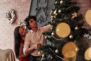 gut gemacht. Romantisches Paar schmückt Weihnachtsbaum im Zimmer mit brauner Wand und Kamin foto