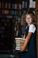 süßes kleines Mädchen mit Brille steht in der Bibliothek voller Bücher. Konzeption von Bildung foto