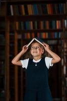 süßes kleines Mädchen mit Brille steht in der Bibliothek voller Bücher. Konzeption von Bildung foto