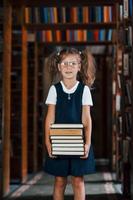 süßes kleines Mädchen mit Brille steht in der Bibliothek voller Bücher. Konzeption von Bildung foto