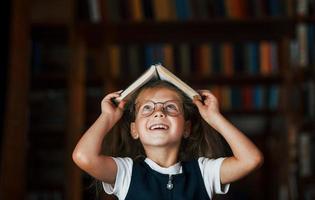 süßes kleines Mädchen mit Brille steht in der Bibliothek voller Bücher. Konzeption von Bildung foto