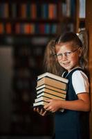 süßes kleines Mädchen mit Brille steht in der Bibliothek voller Bücher. Konzeption von Bildung foto