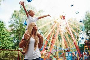 Tochter sitzt auf den Schultern. Fröhliches kleines Mädchen, ihre Mutter, hat eine gute Zeit im Park zusammen in der Nähe von Sehenswürdigkeiten foto