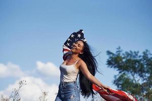 weiblicher patriot läuft mit usa-flagge in den händen draußen im feld gegen blauen himmel foto