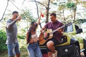 gemeinsam Lieder singen. Freunde haben ein schönes Wochenende im Freien in der Nähe ihres grünen Autos mit Akustikgitarre foto