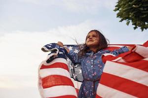 schöner grüner Baum. patriotisches weibliches kind mit amerikanischer flagge in den händen. gegen bewölkten Himmel foto