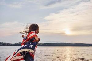 erstaunlicher Sonnenuntergang. patriotisches weibliches kind mit amerikanischer flagge in den händen. gegen bewölkten Himmel foto